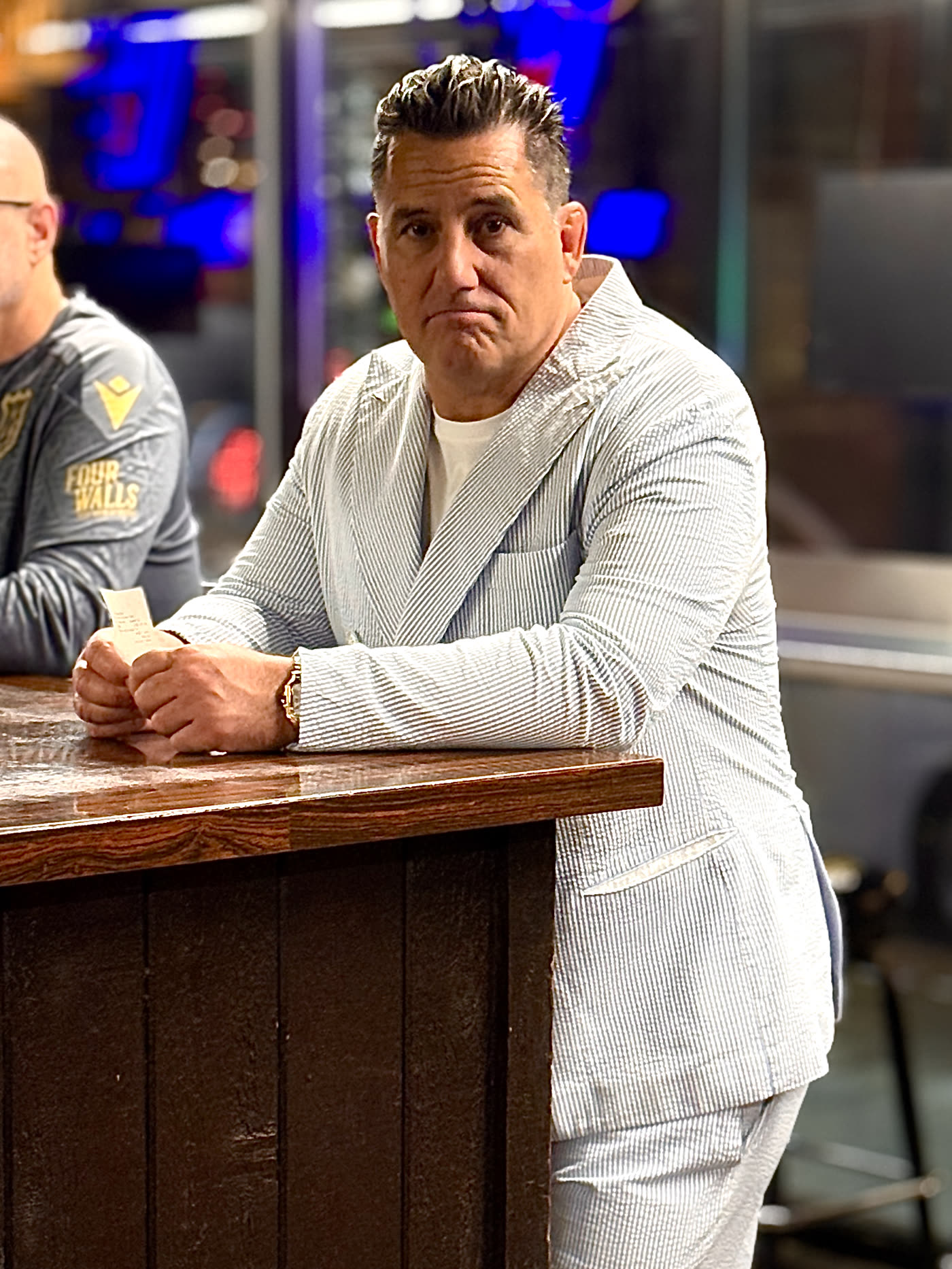 Dino Spencer leaning on a bar in a seersucker jacket, giving a dry, unconvinced look to camera.